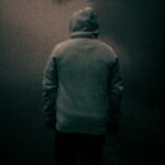 Back view of anonymous male in warm hoodie standing alone in misty forest near trees under gloomy sky