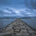 Serene twilight view of a lighthouse guiding ships amidst a rocky coast and cloudy sky.