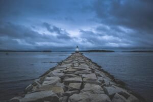 Serene twilight view of a lighthouse guiding ships amidst a rocky coast and cloudy sky.